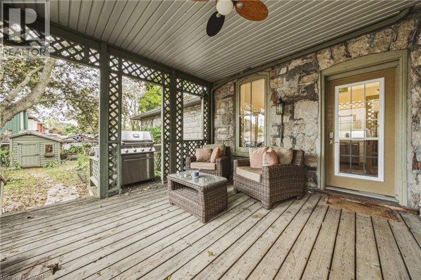 Covered wooden porch with wicker seating, coffee table, gas grill, and view of backyard structures. - 75 Pollock Avenue, Cambridge