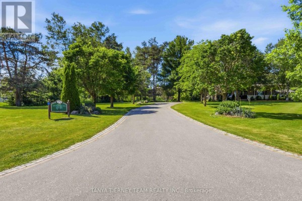 Long paved driveway curving through a manicured lawn with scattered trees and a street sign. - 75 Woodbridge Circle, Scugog