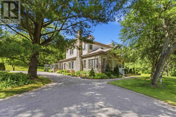 Side view of the property showing a stone chimney, large windows, covered porch, and extensive greenery. - 75 Woodbridge Circle, Scugog