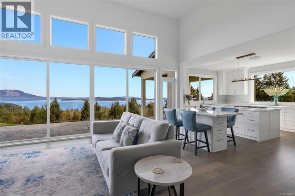 Spacious living room with large windows, grey sofa, and white kitchen island with blue bar stools. - 755 Butterfield Rd, Mill Bay