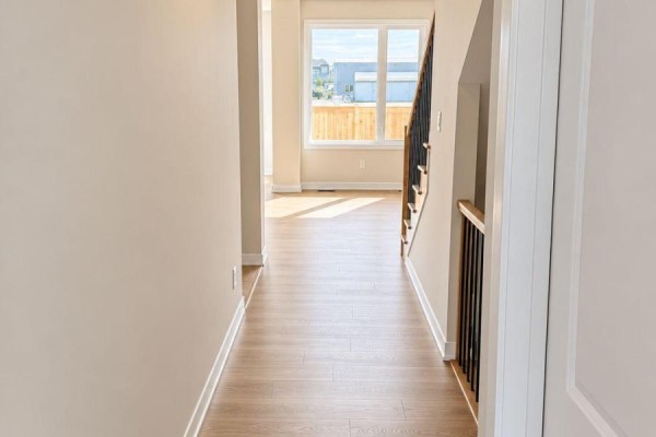 Hallway with light wood floors leading to a living area with large windows and a staircase. - 755 Haflinger Row, Ottawa