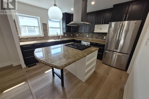 A wide view of a modern kitchen with dark cabinets, granite counters, a stainless steel fridge, and an island with a cooktop. - 757 Pam Crescent, Newmarket