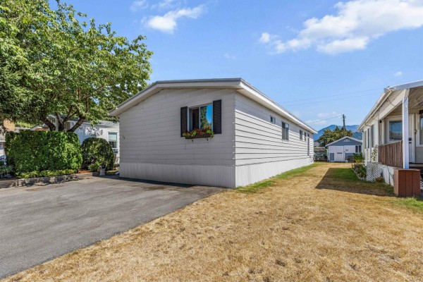View of the mobile home's exterior from the driveway, showing a large tree and neighboring homes. - 76 45640 Watson Road, Garrison Crossing, Chilliwack