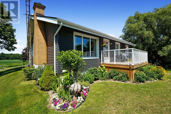 Side view of a house showing dark grey siding, a brick chimney, a wooden deck, and garden beds. - 7638 Telephone Road, Hamilton Township