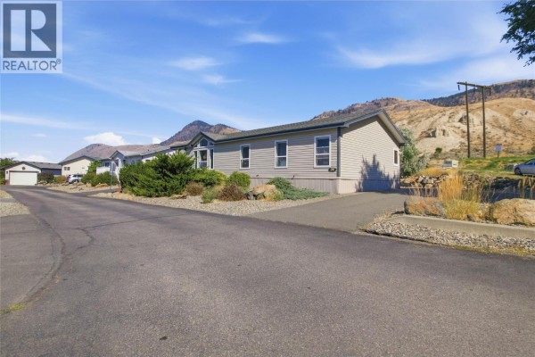 A light gray mobile home with a dark roof is viewed from the street on a sunny day. - 768 Shuswap  E Road Unit# 24, Kamloops