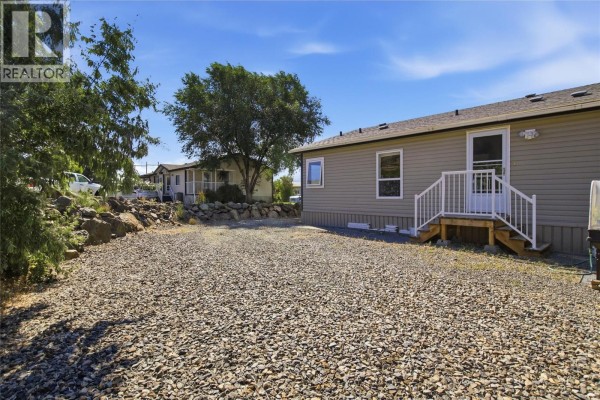 A light gray mobile home with white stairs seen from a gravel yard with trees. - 768 Shuswap  E Road Unit# 24, Kamloops