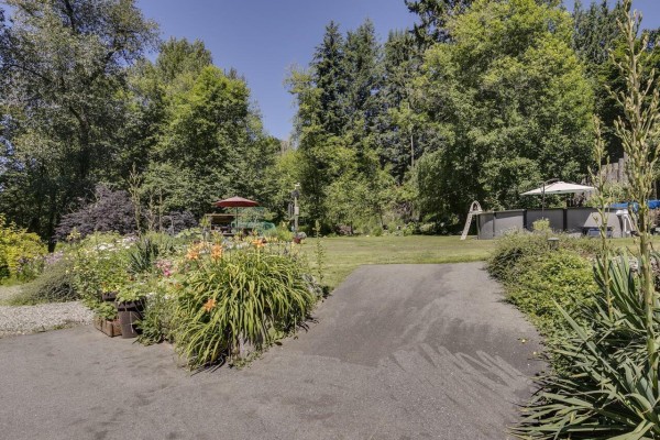 View of the backyard with mature trees, flower beds, and lawn furniture. - 7681 Horne Street, Mission