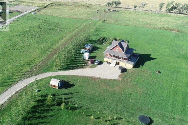 Aerial view of a red house with a wrap-around porch, driveway, and outbuildings in a large grassy field. - 77, 210044 Highway 512, Rural Lethbridge County