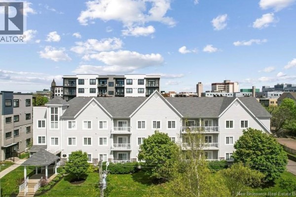 Rear exterior of the light grey condominium building with ground-level access doors and green lawn. - 77 Cameron Street Unit# 305, Moncton