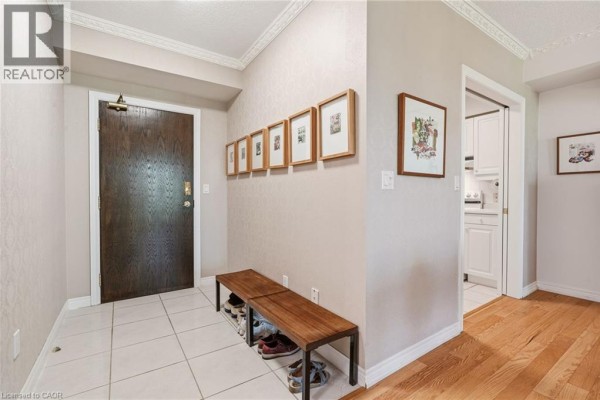 Apartment entryway with a wooden door, a bench, shoes, and framed artwork on wall. - 77 Governor's Road Unit# 404, Dundas