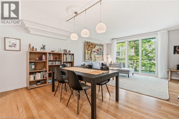 Living and dining area with wood floors, black table, chairs, bookshelf, and large window. - 77 Governor's Road Unit# 404, Dundas