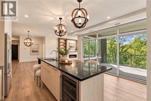 Kitchen island with sink and wine fridge, facing open living area with fireplace and balcony access. - 77 Yates Street Unit# 201, St  Catharines