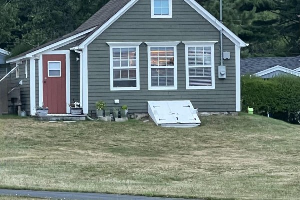 Close-up view of the grey cottage with red front door, white trim windows, and a white basement hatch. - 78 Marriotts Cove Road W, Marriotts Cove
