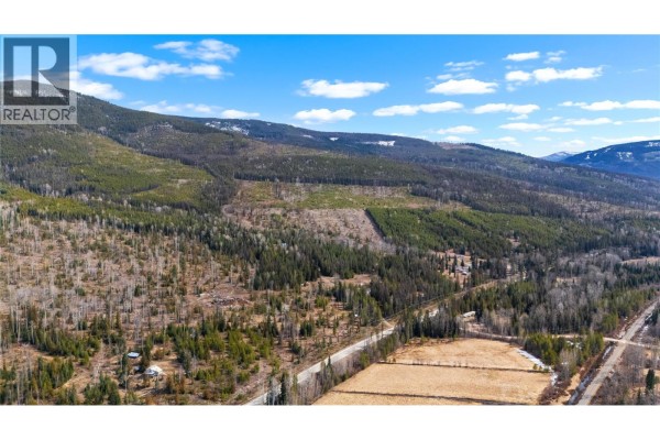Aerial view of a rural property with a paved road running through it, showcasing surrounding hillsides with scattered trees and fields. - 7904 Highway 3, Yahk