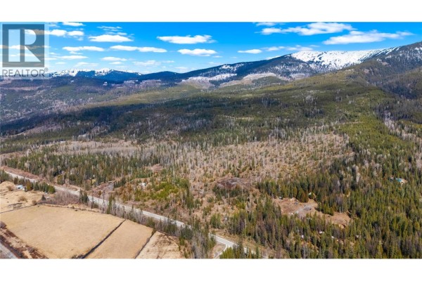 Aerial perspective of a wooded landscape with a paved road, highlighting the varied terrain of hills and distant snow-capped mountains. - 7904 Highway 3, Yahk