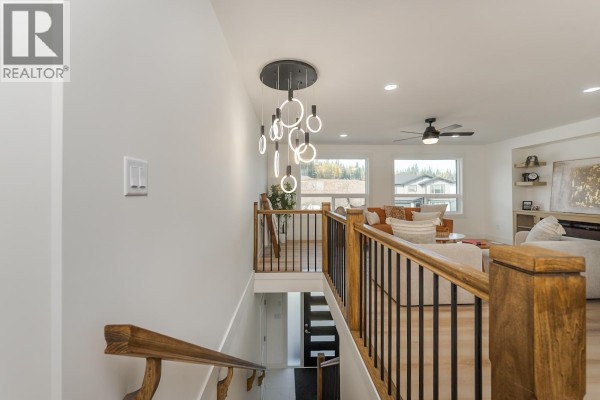 Hallway with wood railings, modern light fixture, and view into a living room with large windows. - 7990 Southridge Avenue, Prince George
