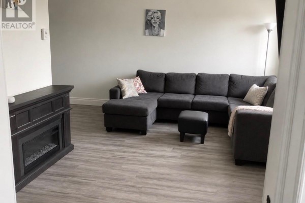 Living room showcasing a large grey sectional sofa, electric fireplace, and light grey wood-look flooring. - 8 Banting Avenue, Wabush