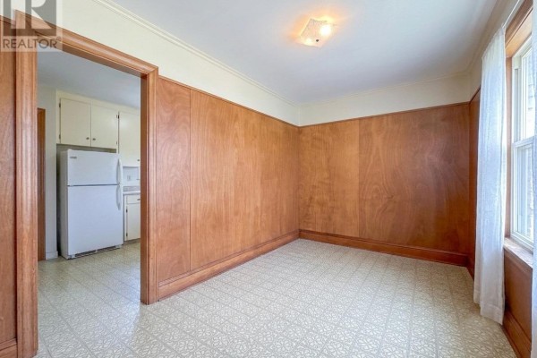 Interior room with wood-paneled walls, beige patterned floor, and view into a kitchen with white appliances. - 8 Falconwood Drive, Charlottetown