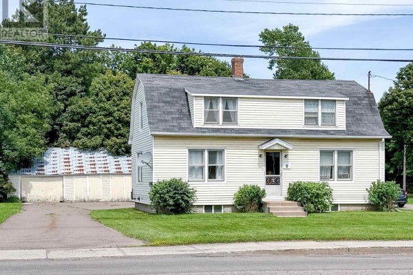 Side view of a two-story cream-colored house with a driveway, asphalt road, and mature trees. - 8 Falconwood Drive, Charlottetown