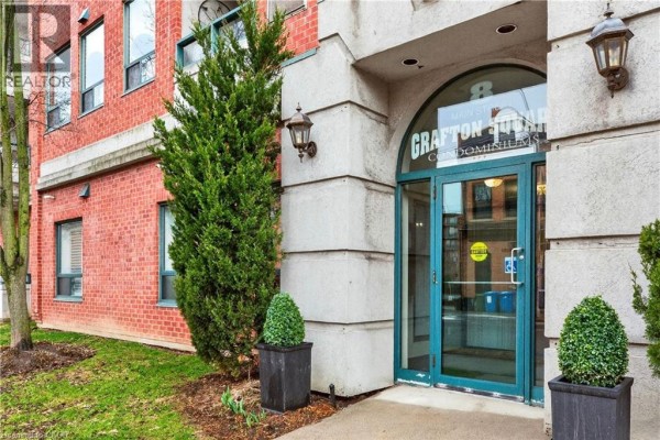 Detail of the apartment building entrance with a large evergreen tree and planter flanking the glass doors. - 8 Main Street Unit# 101, Dundas