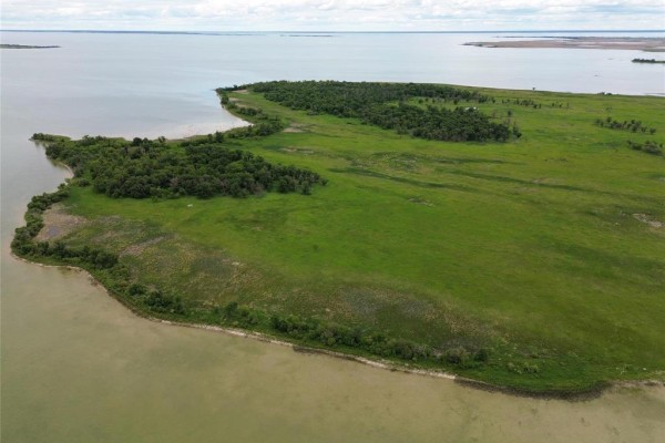 Aerial perspective of a vast, open green field with trees along the edges and water in the foreground. - 8 Reykjavik Road, Lake Manitoba Narrows