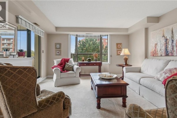 Living room with beige carpet, white sofa, and a bay window overlooking trees. - 8 Village Green Boulevard Unit# 510, Hamilton