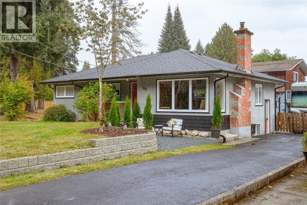Front corner view of a grey bungalow with grey shingle siding, brick accents, and a paved driveway. - 80 North Shore Rd, Lake Cowichan