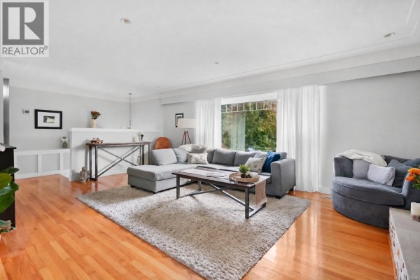 Living room with a large sectional sofa, wood floors, and a window with greenery view. - 800 Alvarado Terr, Saanich