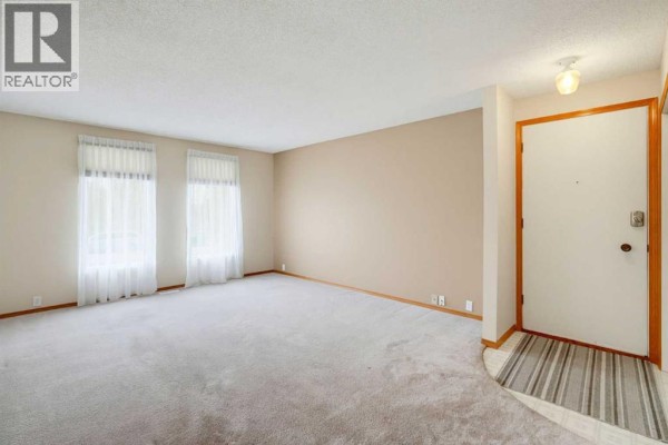 Living room with light beige carpet, beige walls, and two large windows with sheer curtains. - 8007 Huntington Street Ne, Calgary