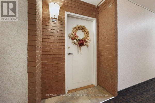 Hallway outside apartment 801 with a white door, brick accent walls, and a decorative wreath. - 801 - 1966 Main Street W, Hamilton