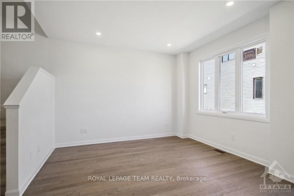 Empty room with white walls, hardwood floors, and a large window overlooking houses. - 805 Everbloom Lane, Ottawa