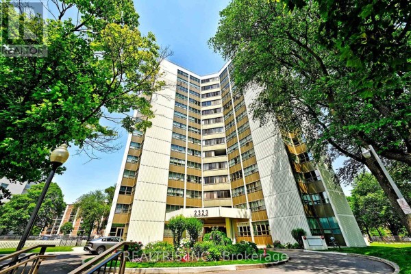 View from below of a tall apartment building with trees partially obscuring the facade. - 807 - 2323 Confederation Parkway, Mississauga