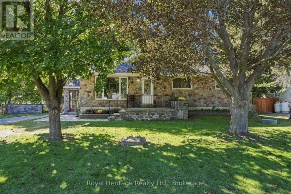 Front of the bungalow framed by mature trees and a green lawn. - 8074 Tinkerville Road, Port Hope