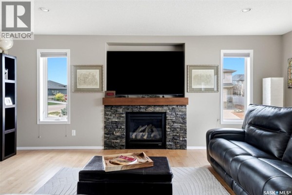 Close-up of the living room stone fireplace with a wooden mantel, surrounded by framed art and windows. - 8097 Wascana Gardens Crescent, Regina