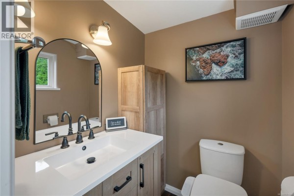 Bathroom with vanity, sink, arched mirror, and toilet. Wood-look cabinet on right. - 81 5250 Beaver Harbour Rd, Port Hardy