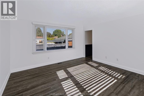 Another view of the empty bedroom with white walls, dark vinyl flooring, and a large window with blinds. - 81 Devon Road, Chatham