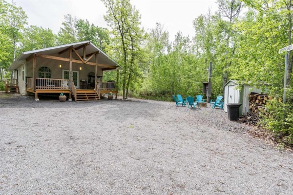 Exterior view of a light beige bungalow with a covered porch, gravel driveway, patio chairs, and a fire pit area. - 81 Reisswood Drive, Traverse Bay