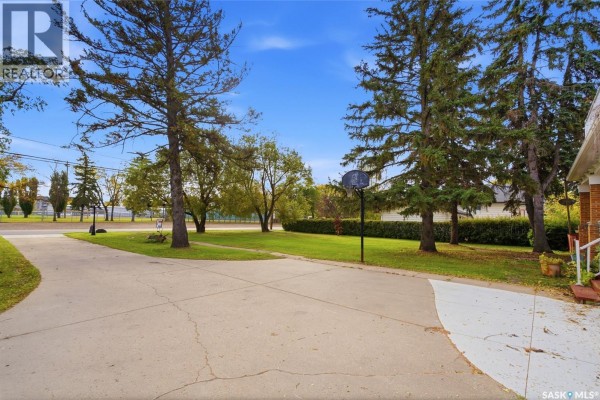 Concrete driveway with a basketball hoop in a grassy yard with mature trees. - 810 4th Avenue, Rosthern