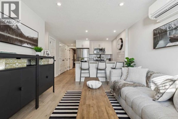 Open concept living and kitchen area with grey sofa, island, bar stools, and stainless steel appliances. - 812, 135 Mahognay Parade Se, Calgary