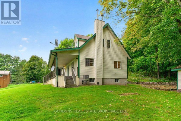 Side view of the house showing a covered deck, white siding, and a large stone chimney. - 813 Pierce's Lane, Leeds And The Thousand Islands