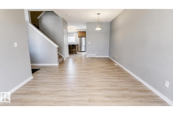 Empty living and dining room with light wood laminate flooring and a view into the kitchen. - 8145 Chappelle Wy Sw, Edmonton