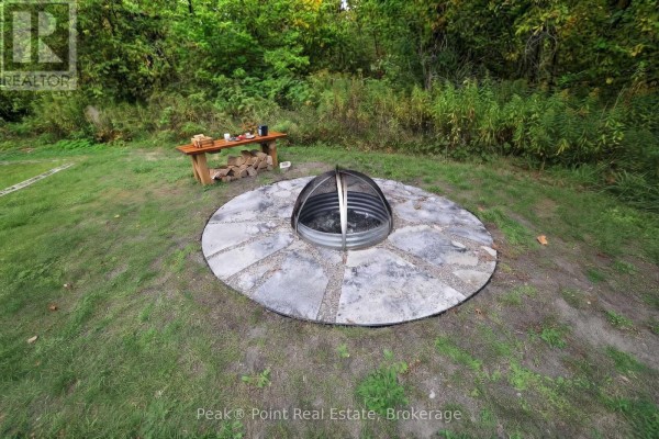 Outdoor circular stone fire pit with a metal grate and a wooden bench with firewood nearby. - 815 Main Street, South Bruce Peninsula