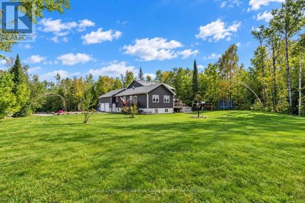 Rear exterior of a dark-sided house with a large green lawn and trees. - 819 County 49 Road, Kawartha Lakes