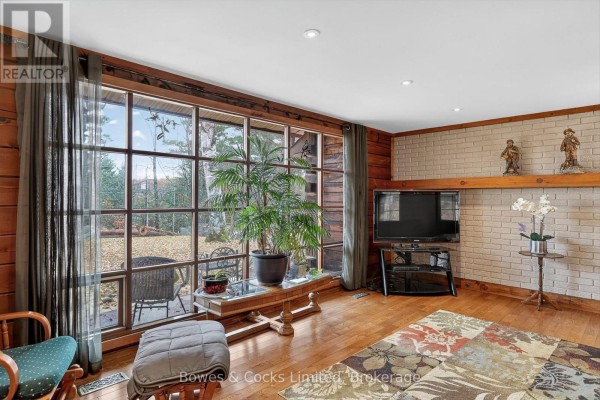 Living room with large window, wood paneling, and brick accent wall. - 82 Bay Lake Road, Bancroft