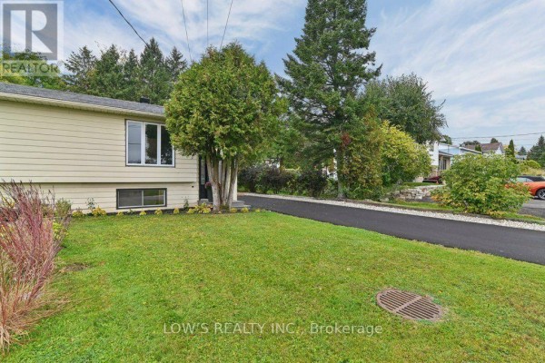 Front lawn and driveway of a beige house with mature trees and lush green grass. - 82 Bertha Street, Champlain