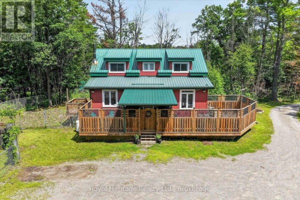 Red cottage with green roof and wooden deck surrounded by trees. - 82 Kennedy Drive, Trent Lakes