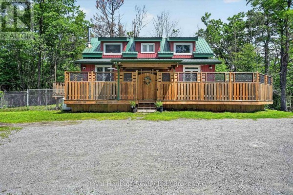 Front view of a red cottage with a wooden deck and gravel driveway. - 82 Kennedy Drive, Trent Lakes