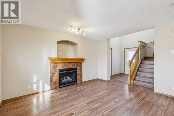 Living room with a gas fireplace, wooden mantel, and hardwood floors, with stairs visible in the background. - 8226 Saddleridge Drive Ne, Calgary