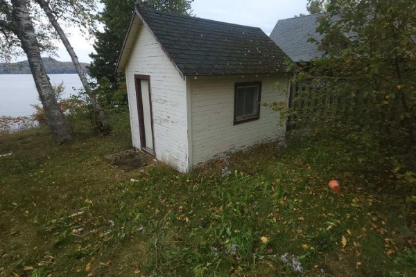 A small, white shed with peeling paint and a brown door sits near a lake. - 83 Fell Rd, R.r. #2 Baptiste, Hastings Highlands
