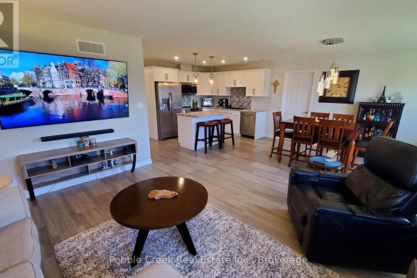 Open concept living area with a large TV, fireplace, dining table, and kitchen in the background. - 83 Huron Heights Drive, Ashfield Colborne Wawanosh
